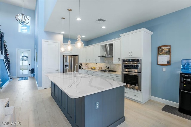 kitchen with wall chimney exhaust hood, stainless steel appliances, a large island with sink, decorative light fixtures, and white cabinetry