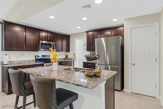 kitchen with light stone counters, a kitchen bar, a center island with sink, light tile patterned flooring, and appliances with stainless steel finishes