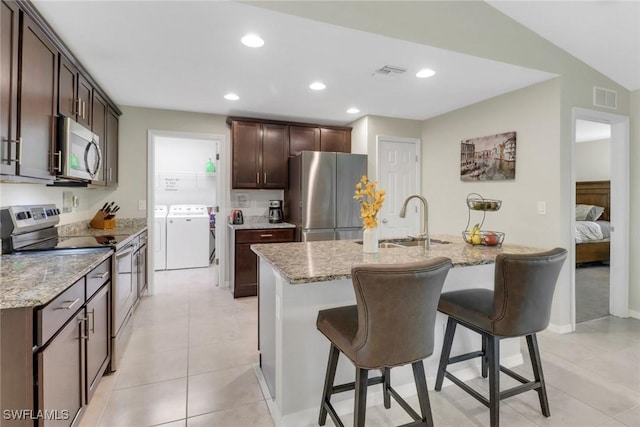 kitchen with washer and clothes dryer, a center island with sink, sink, light stone countertops, and appliances with stainless steel finishes