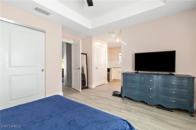 bedroom featuring light wood-type flooring, a tray ceiling, ensuite bath, and ceiling fan