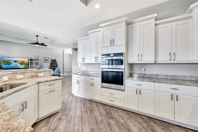 kitchen with ceiling fan, double oven, light hardwood / wood-style floors, light stone counters, and white cabinetry