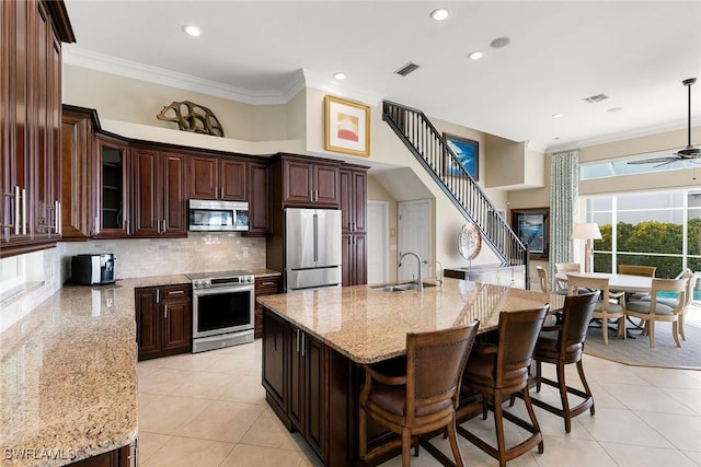 kitchen featuring dark brown cabinetry, ceiling fan, stainless steel appliances, an island with sink, and ornamental molding