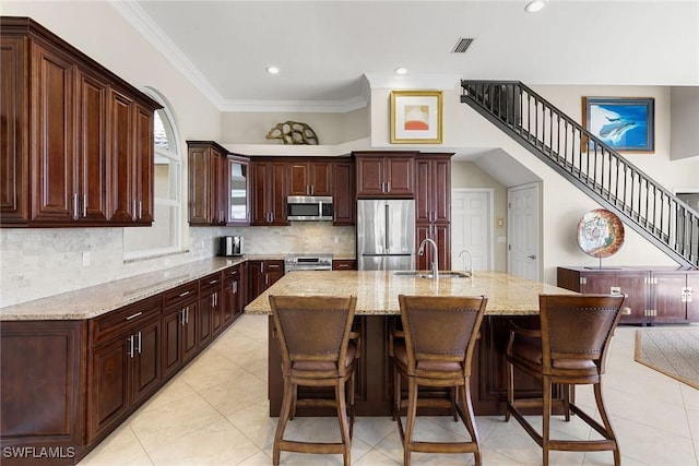 kitchen with light stone counters, stainless steel appliances, a kitchen island with sink, crown molding, and sink