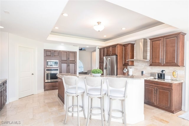 kitchen with wall chimney exhaust hood, a kitchen island with sink, a tray ceiling, decorative backsplash, and appliances with stainless steel finishes