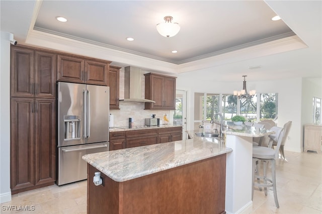 kitchen with high end fridge, a tray ceiling, sink, wall chimney range hood, and an island with sink