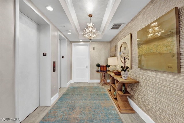 corridor featuring elevator, a tray ceiling, a chandelier, and light hardwood / wood-style flooring