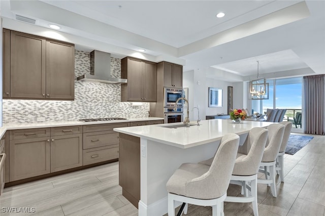 kitchen with appliances with stainless steel finishes, backsplash, an island with sink, a raised ceiling, and wall chimney exhaust hood