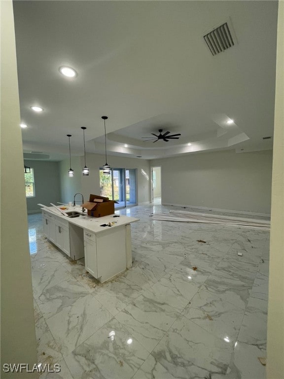 kitchen featuring a raised ceiling, a kitchen island with sink, ceiling fan, white cabinetry, and hanging light fixtures