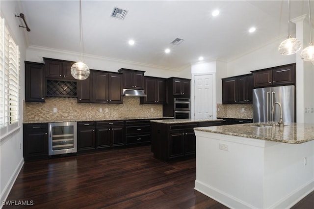 kitchen featuring appliances with stainless steel finishes, a kitchen island with sink, pendant lighting, dark hardwood / wood-style floors, and wine cooler