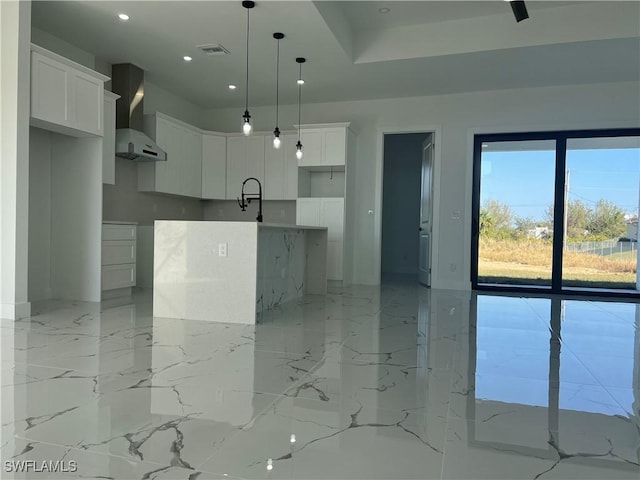 kitchen featuring white cabinets, wall chimney exhaust hood, an island with sink, and hanging light fixtures