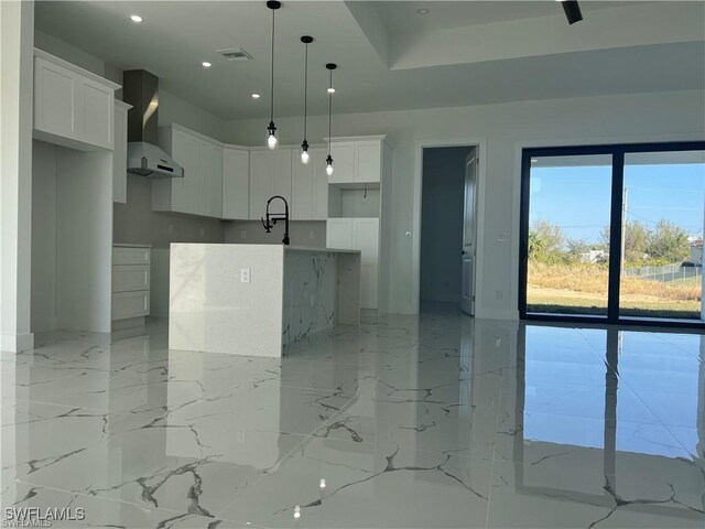 kitchen with white cabinetry, marble finish floor, wall chimney range hood, an island with sink, and pendant lighting