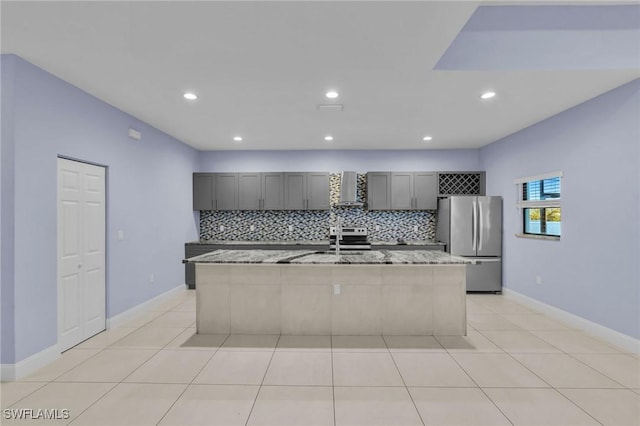 kitchen featuring gray cabinetry, wall chimney range hood, light stone counters, a center island with sink, and appliances with stainless steel finishes