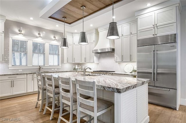 kitchen featuring wall chimney range hood, pendant lighting, a kitchen island with sink, appliances with stainless steel finishes, and light wood-type flooring