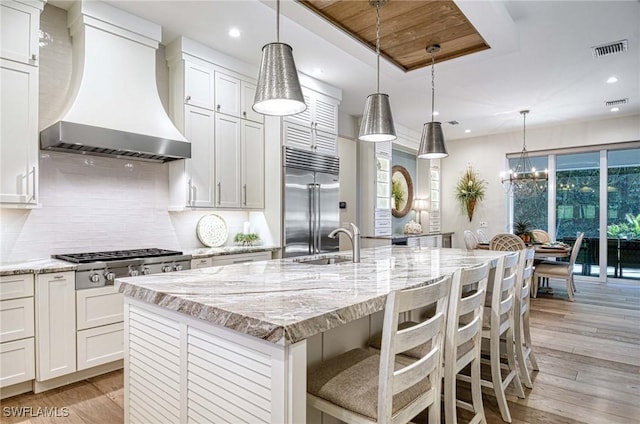 kitchen with wall chimney range hood, light wood-type flooring, an island with sink, white cabinetry, and stainless steel appliances