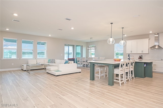 kitchen with pendant lighting, white cabinets, wall chimney range hood, light hardwood / wood-style flooring, and an island with sink