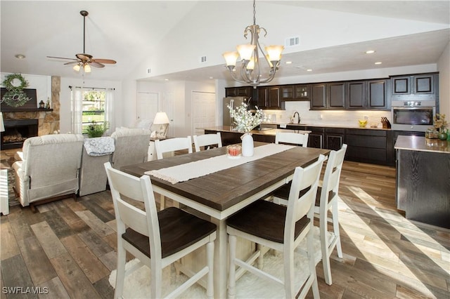 dining space featuring hardwood / wood-style flooring, a stone fireplace, high vaulted ceiling, and ceiling fan with notable chandelier