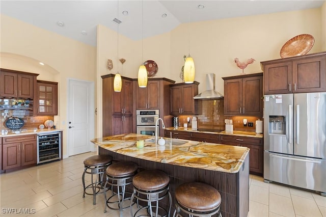 kitchen with wine cooler, appliances with stainless steel finishes, backsplash, wall chimney range hood, and an island with sink