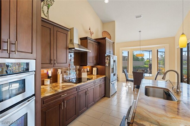 kitchen with hanging light fixtures, sink, light stone countertops, wall chimney range hood, and stainless steel appliances