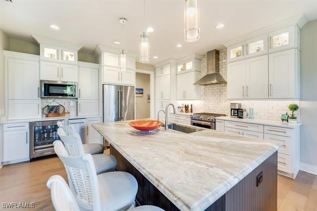 kitchen featuring white cabinetry, sink, stainless steel appliances, wall chimney range hood, and an island with sink