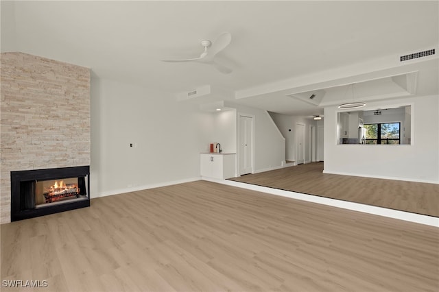 unfurnished living room featuring light hardwood / wood-style flooring, a stone fireplace, ceiling fan, and a tray ceiling