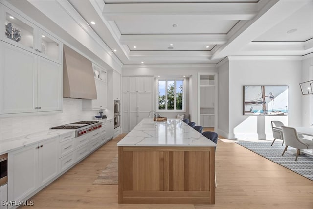 kitchen with stainless steel gas stovetop, a center island with sink, wall chimney exhaust hood, light stone countertops, and white cabinetry