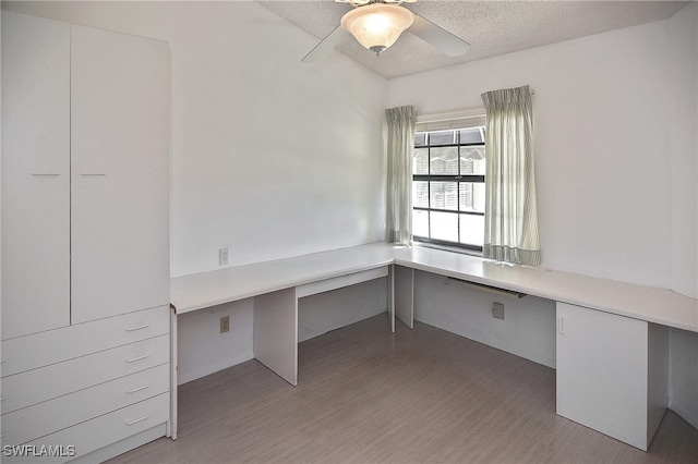 unfurnished office featuring ceiling fan, light hardwood / wood-style floors, built in desk, and a textured ceiling