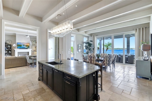 kitchen featuring light stone countertops, sink, a water view, a center island with sink, and beamed ceiling