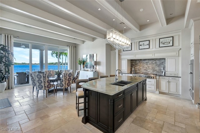 kitchen with sink, light stone counters, beamed ceiling, an island with sink, and a water view