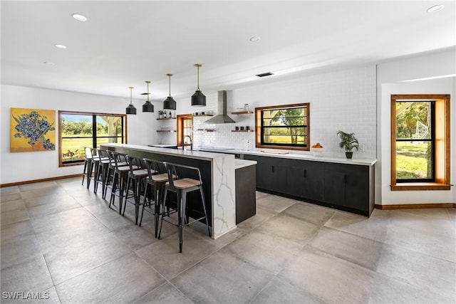 kitchen featuring a kitchen island with sink, a kitchen breakfast bar, wall chimney range hood, decorative backsplash, and open shelves