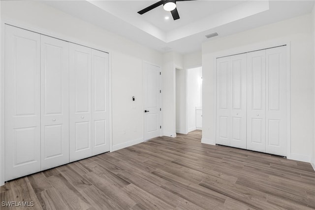 unfurnished bedroom featuring ceiling fan, multiple closets, light wood-type flooring, and a tray ceiling