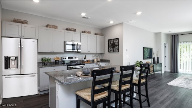 kitchen featuring a kitchen island with sink, stone counters, dark wood-type flooring, a kitchen breakfast bar, and appliances with stainless steel finishes