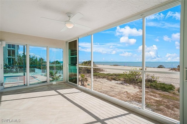 unfurnished sunroom with a view of the beach, a water view, ceiling fan, and a healthy amount of sunlight