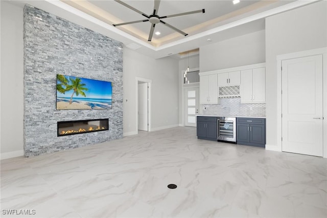 unfurnished living room with ceiling fan, beverage cooler, a high ceiling, a raised ceiling, and a stone fireplace