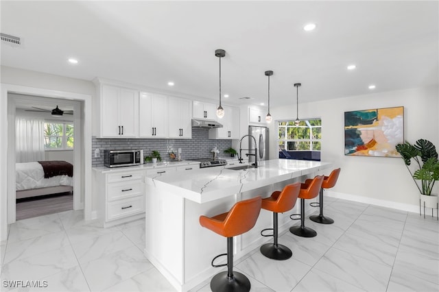 kitchen featuring appliances with stainless steel finishes, a kitchen bar, white cabinetry, hanging light fixtures, and a kitchen island with sink