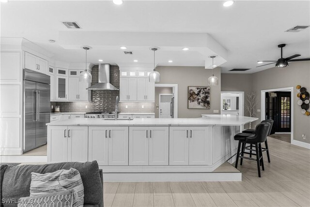 kitchen with a center island with sink, glass insert cabinets, light countertops, wall chimney range hood, and built in fridge