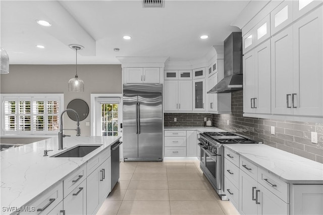 kitchen with decorative light fixtures, high end appliances, white cabinetry, a sink, and wall chimney exhaust hood