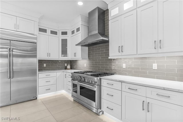 kitchen with light stone counters, white cabinets, wall chimney exhaust hood, high end appliances, and glass insert cabinets