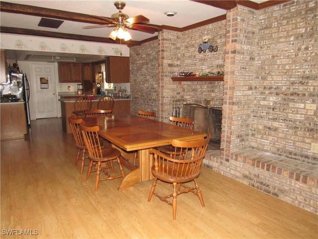 dining room with beam ceiling, ceiling fan, a brick fireplace, brick wall, and light wood-type flooring