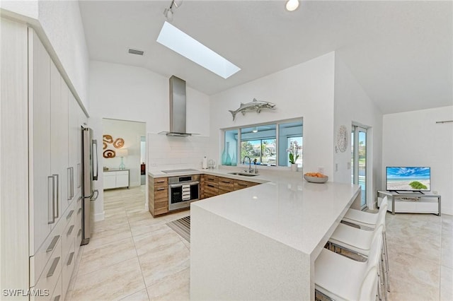 kitchen featuring wall chimney range hood, a skylight, light tile patterned floors, a kitchen bar, and stainless steel appliances