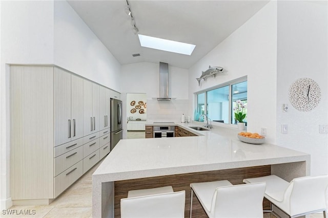 kitchen featuring kitchen peninsula, appliances with stainless steel finishes, wall chimney exhaust hood, sink, and a breakfast bar area