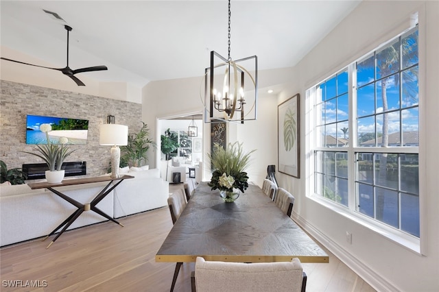 dining room with ceiling fan with notable chandelier, light wood-type flooring, and a stone fireplace