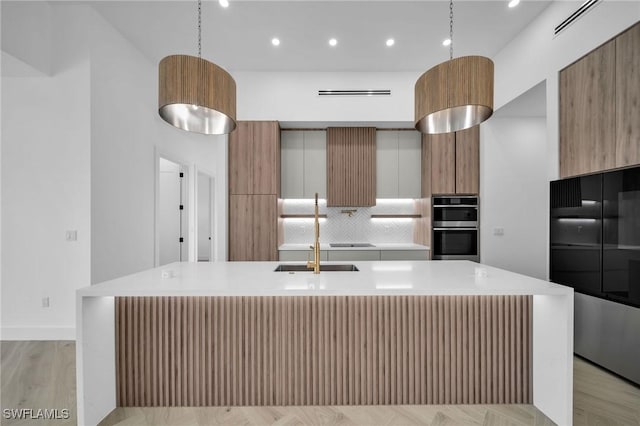 kitchen featuring decorative backsplash, double oven, decorative light fixtures, light hardwood / wood-style flooring, and a center island with sink