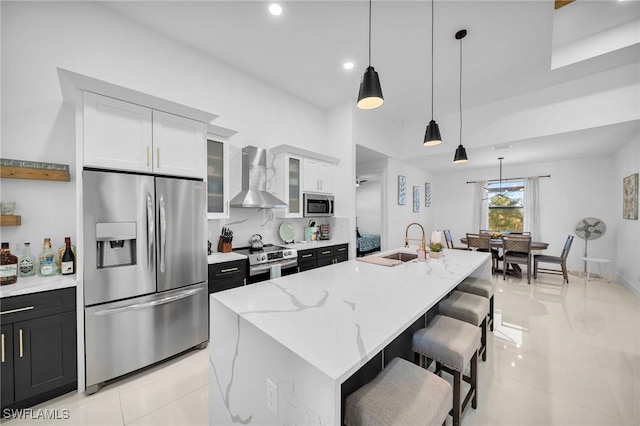 kitchen with a center island with sink, white cabinets, wall chimney range hood, appliances with stainless steel finishes, and decorative light fixtures