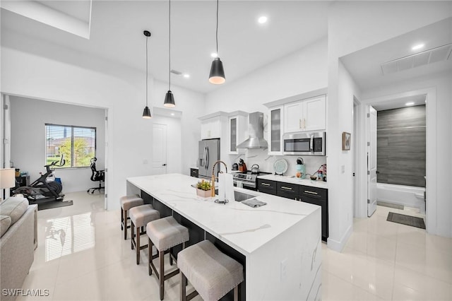 kitchen featuring a center island with sink, wall chimney range hood, decorative light fixtures, white cabinetry, and stainless steel appliances