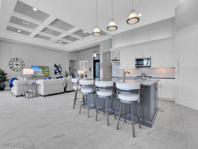 kitchen with a kitchen bar, coffered ceiling, a kitchen island with sink, decorative light fixtures, and white cabinetry