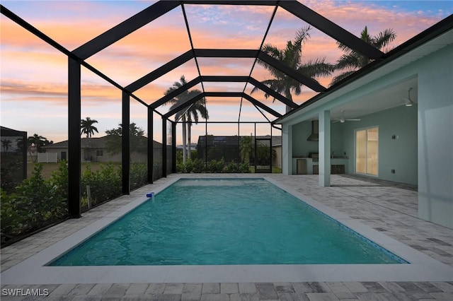 pool at dusk with glass enclosure, ceiling fan, exterior kitchen, and a patio