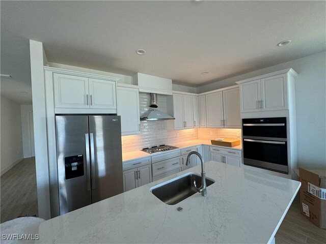 kitchen featuring light stone countertops, appliances with stainless steel finishes, sink, wall chimney range hood, and white cabinets