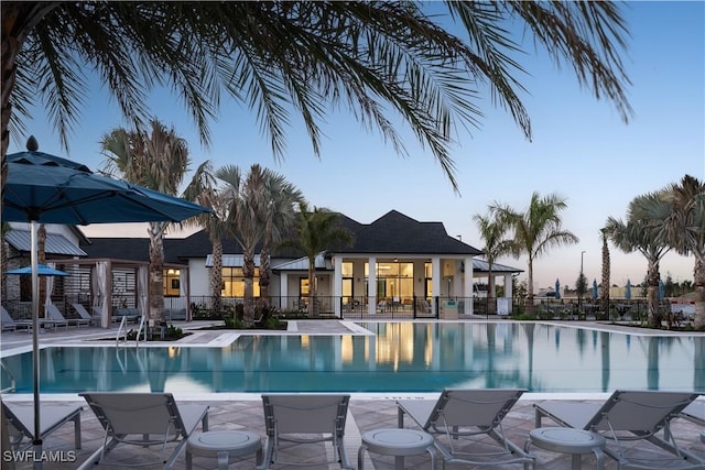 pool at dusk with a patio area, fence, and a community pool