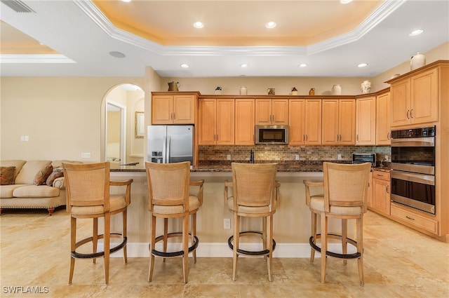 kitchen with stainless steel appliances, a raised ceiling, dark stone countertops, a kitchen island with sink, and a breakfast bar