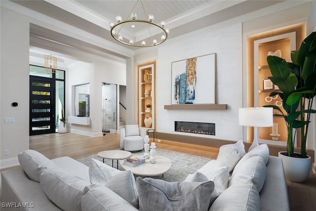 living room featuring a large fireplace, a raised ceiling, built in features, a notable chandelier, and light wood-type flooring